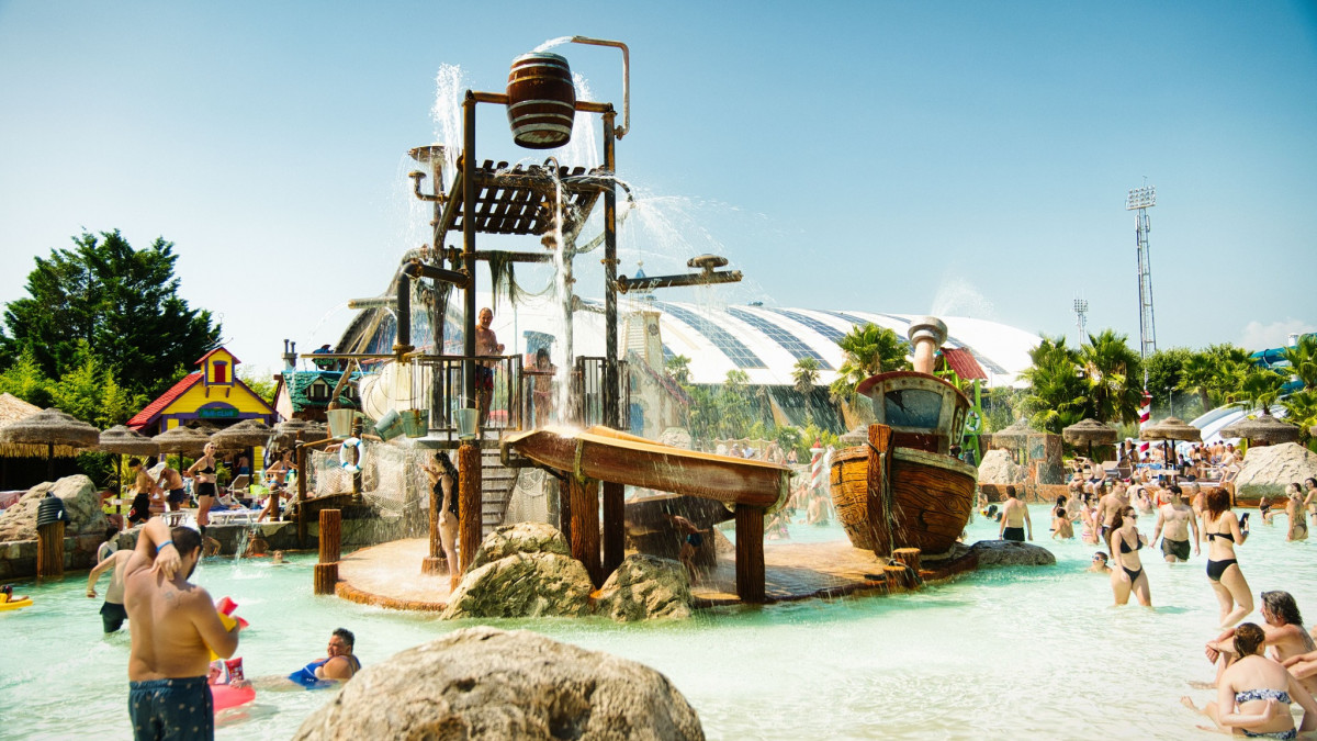 Children enjoying a pirate ship water playground with tipping buckets in a shallow pool, crowded with families having fun.