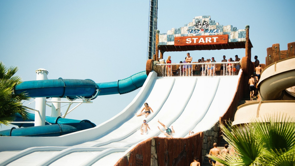 Water slide “Fast'n Race” with children sliding down while others wait at the top; a blue enclosed water slide is visible next to the starting platform.