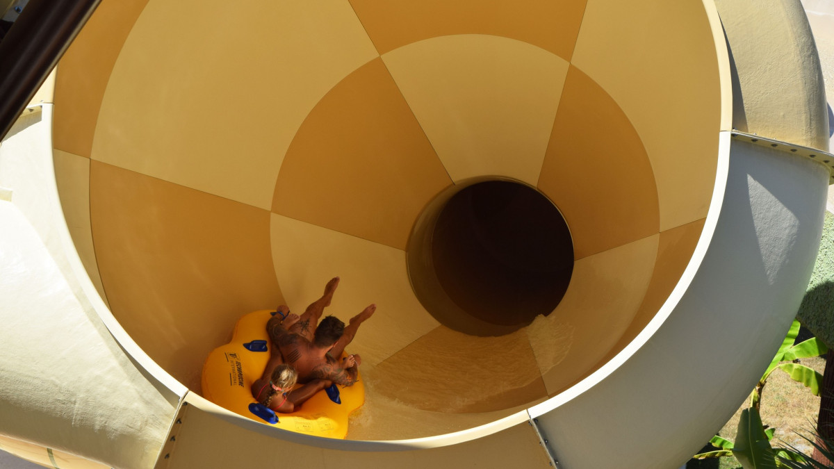 Father and daughter seen from behind on a two-seat inflatable raft, about to slide into the yellow Vortex funnel water slide