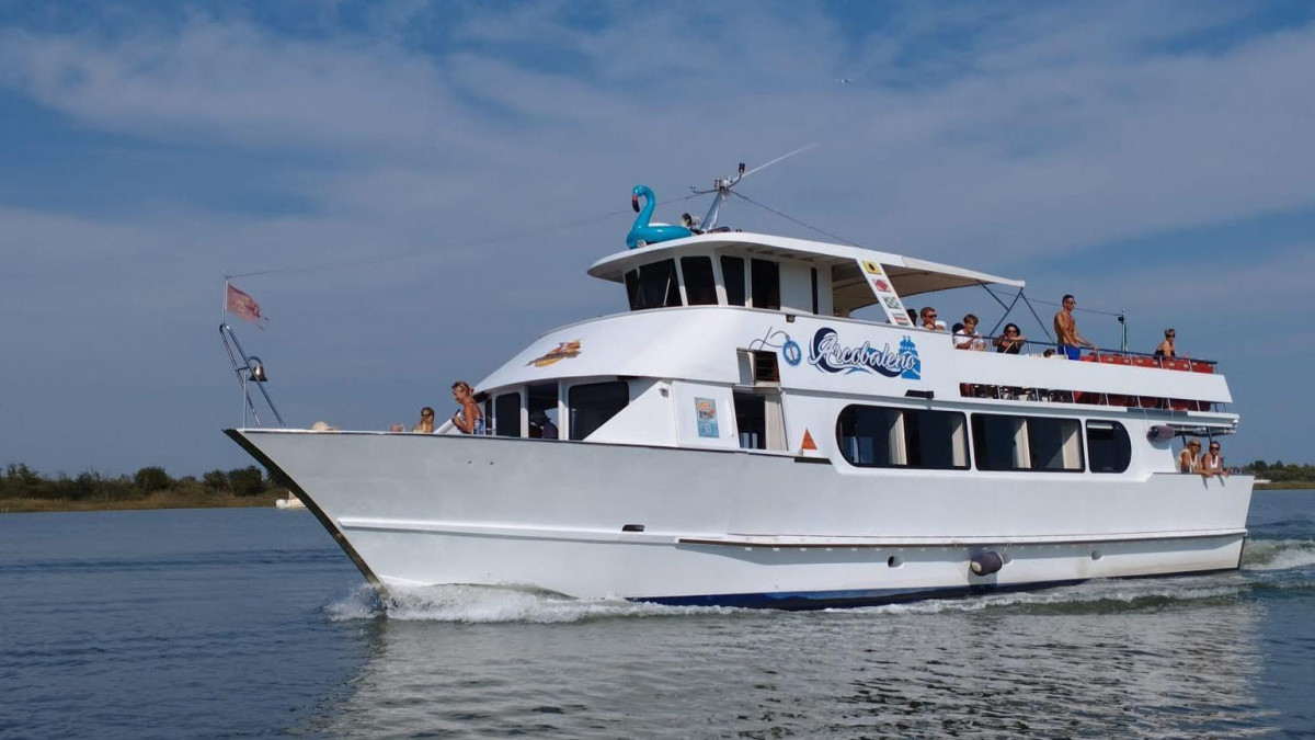 white Arcobaleno tour boat cruising through the blue waters of the Caorle Lagoon under a clear sky, with passengers enjoying the upper deck