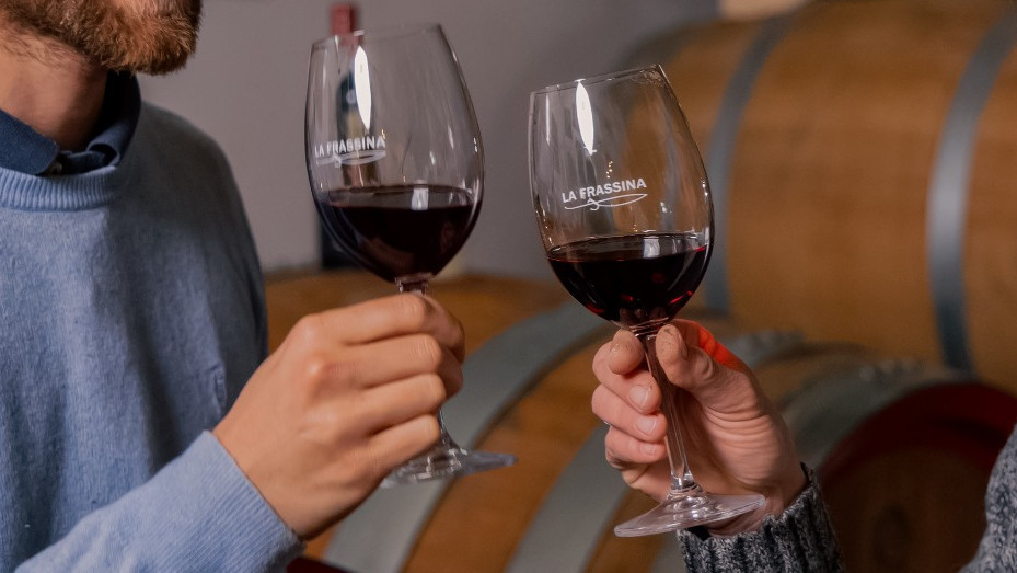 Two people toasting with glasses of red wine labeled "La Frassina" in a cellar with wooden wine barrels in the background.
