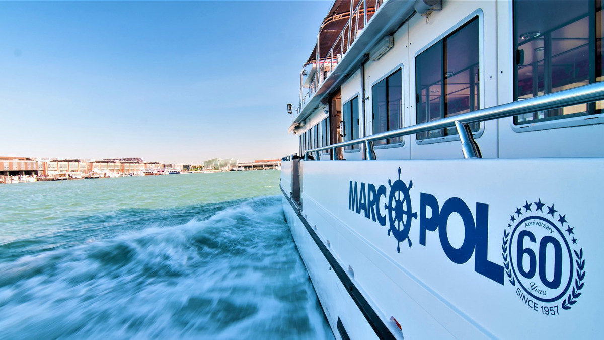 Side view of the white Marco Polo tour boat cruising on the water, showing the 60th-anniversary logo and passenger windows under a clear blue sky.