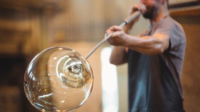Artisan glassblower blowing air through a long metal pipe into a large, glowing glass sphere during a demonstration of traditional Venetian glass blowing
