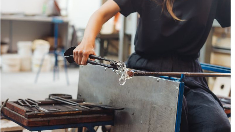 An artisan woman using metal tools to shape molten glass on a blowpipe inside a traditional Venetian glassmaking workshop