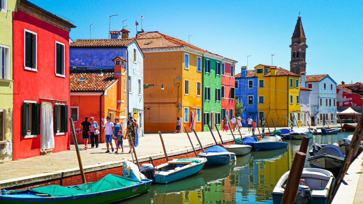 Row of brightly colored houses in red, yellow, and blue along a canal with moored boats and a leaning bell tower in Burano, Venice.