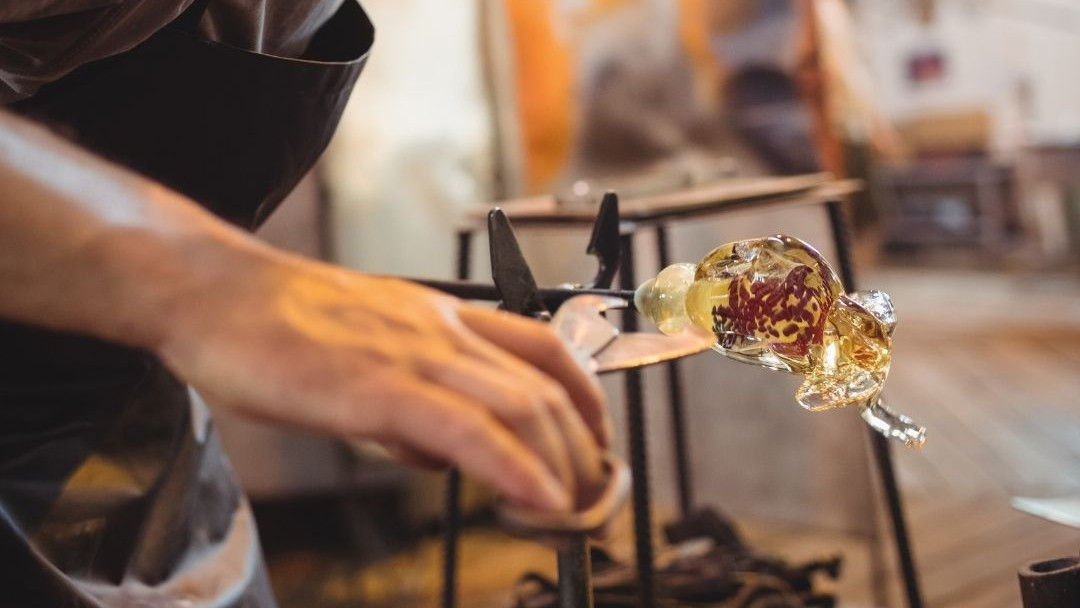 Close-up of a glassmaker's hands using metal jacks to skillfully shape a piece of glowing, molten glass in a traditional furnace workshop
