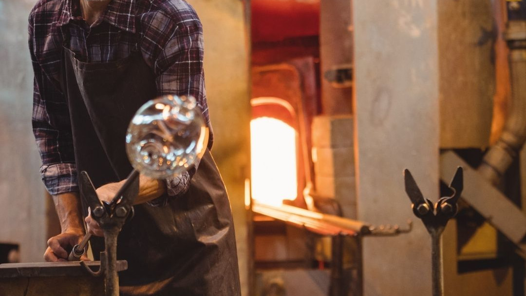 Professional glassblower shaping molten glass with traditional tools in a Murano workshop with a glowing furnace in the background