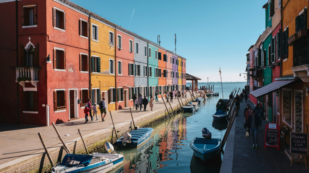 Sunlit canal walk in Burano, Venice, featuring bright multi-colored houses, docked small boats, and local restaurants under a clear blue sky.