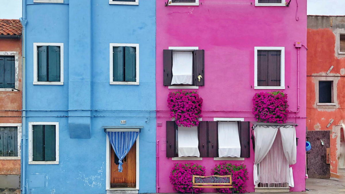 Close-up of vibrant blue and pink houses in Burano, Venice, featuring flower-filled window boxes, dark shutters, and a traditional striped door curtain.