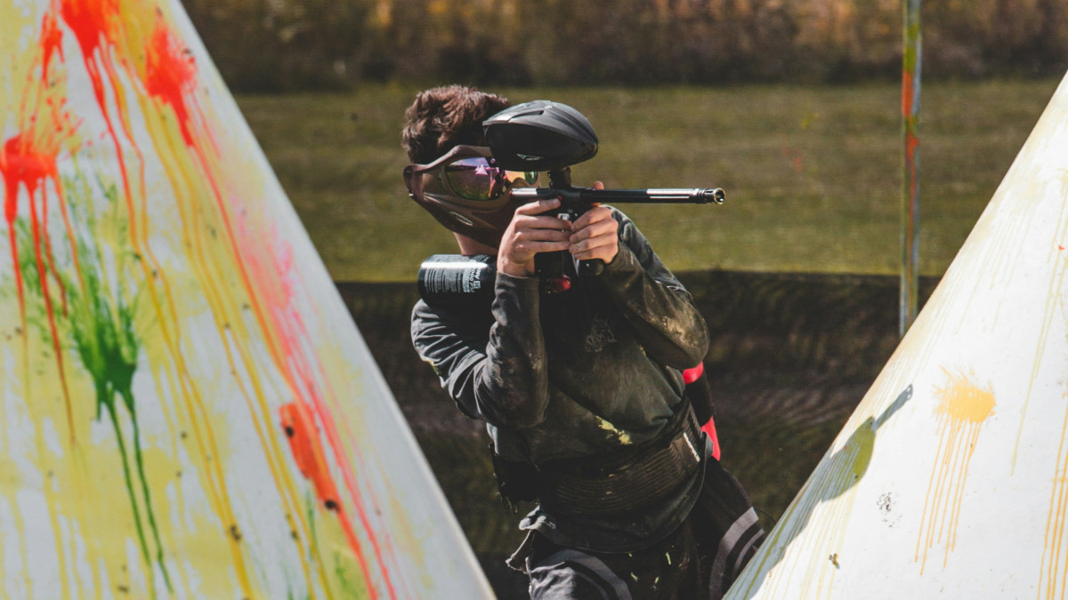 Paintball player in tactical gear taking cover behind a paint-splattered inflatable bunker while aiming a marker during a competitive outdoor match