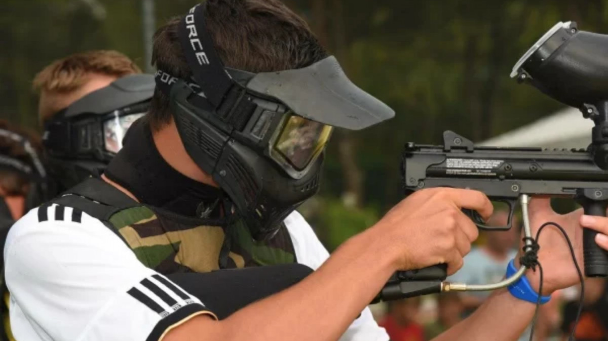 Action shot of a paintball player in a tactical mask and camouflage vest aiming a marker during an outdoor tournament match