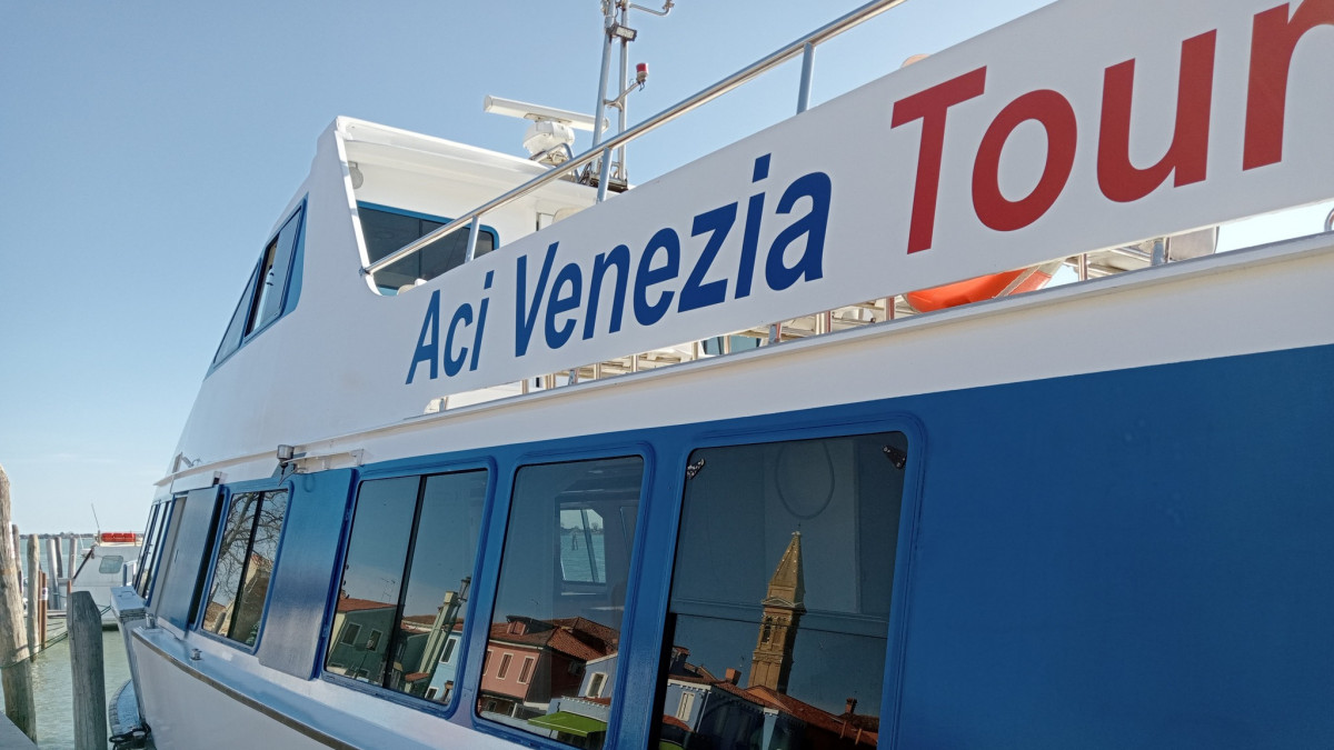 Close-up of the AciVenezia Tourist logo on a white and blue boat docked in the Venetian lagoon with city reflections in the windows