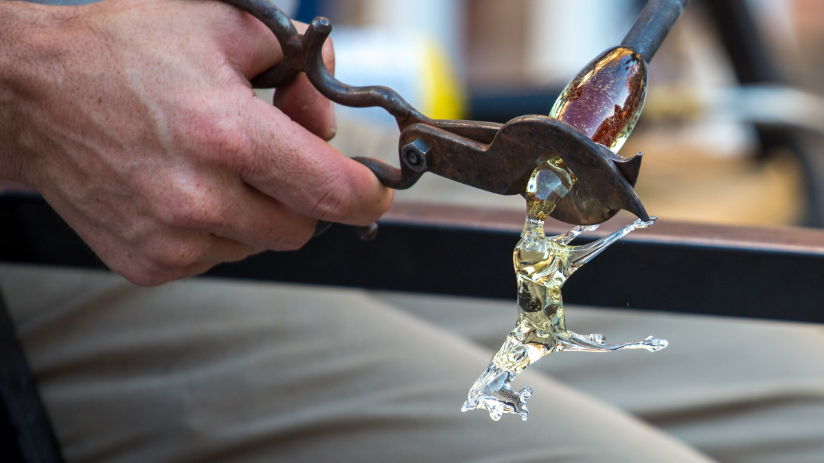 Close-up of a glassblower using metal shears to shape a glowing, molten glass figurine during a traditional glassmaking process