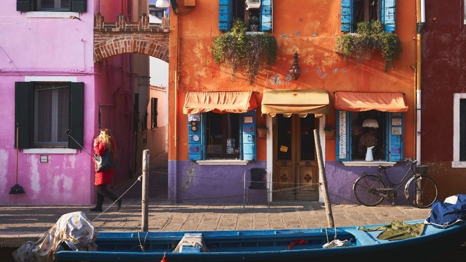 Colorful pink and orange houses in Burano, Venice, with a blue boat in the foreground and a woman walking along the canal