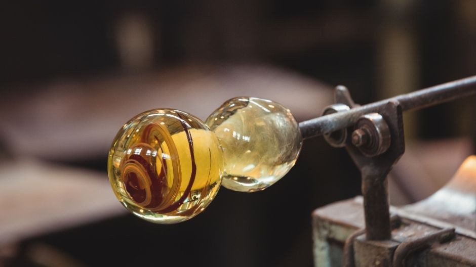 Close-up of a hand-blown glass sphere with yellow and brown swirls being shaped on a metal rod in a glassmaker furnace