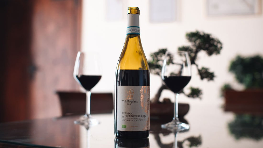 A bottle of Villa Bogdano Refosco dal Peduncolo Rosso Lison-Pramaggiore DOC organic red wine on a table with two glasses.