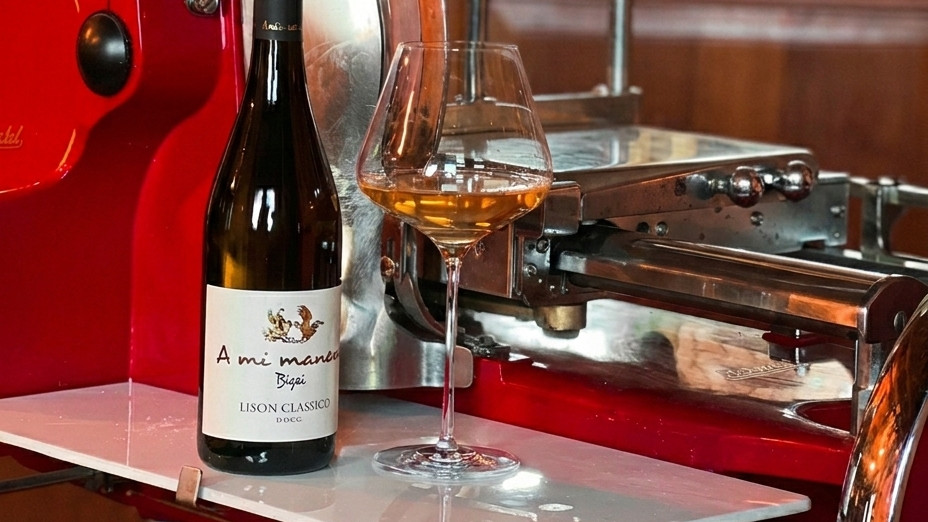 A Mi Manera "Bigai" Lison Classico DOCG white wine bottle and glass in front of a vintage red meat slicer at Cantina A Mi Manera, Portogruaro.