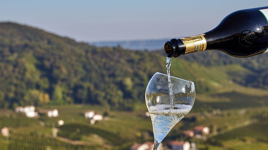 Chilled Prosecco Superiore sparkling wine being poured in a glass with a scenic view of the Valdobbiadene vineyard hills in the background.