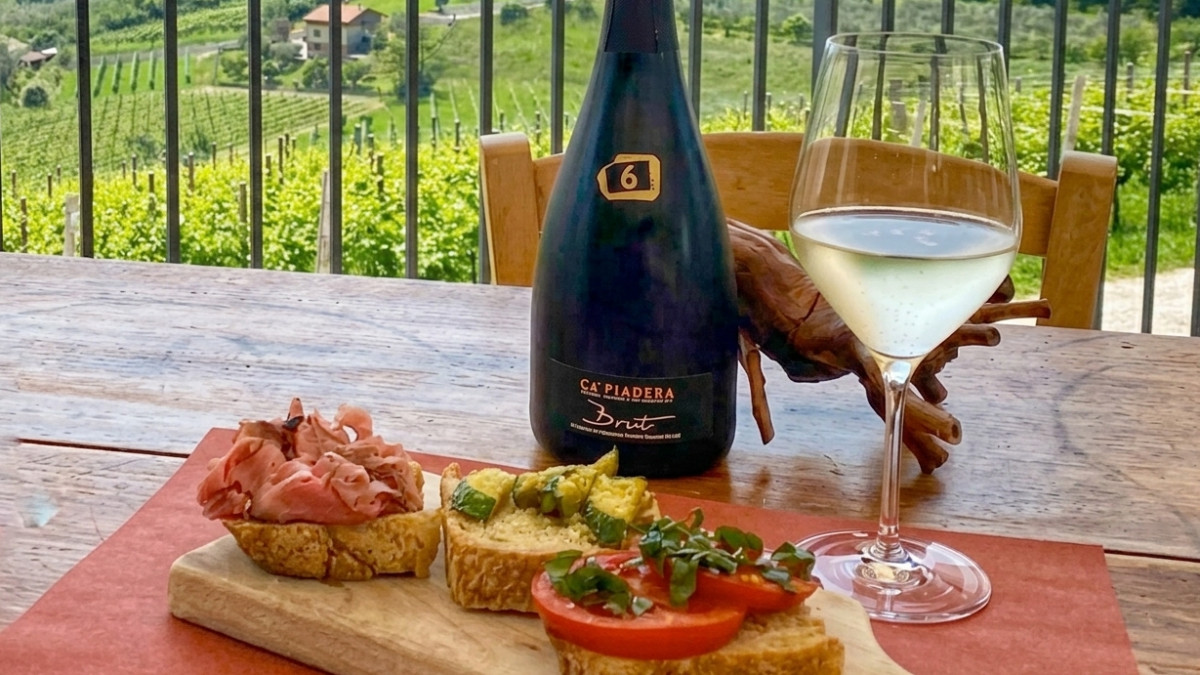 Bottle of Ca' Piadera Brut Prosecco with a glass of white wine and Italian bruschetta appetizers on a wooden table overlooking vineyards.