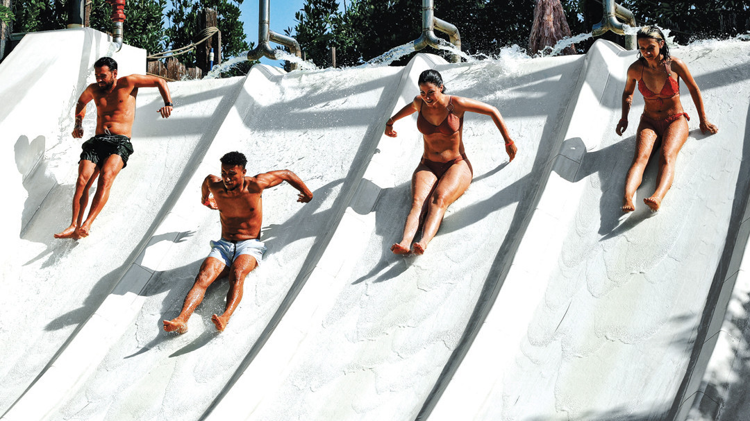 Four friends racing down a multi-lane white water slide at Caneva Acquapark, Italy, splashing through the water on a sunny day