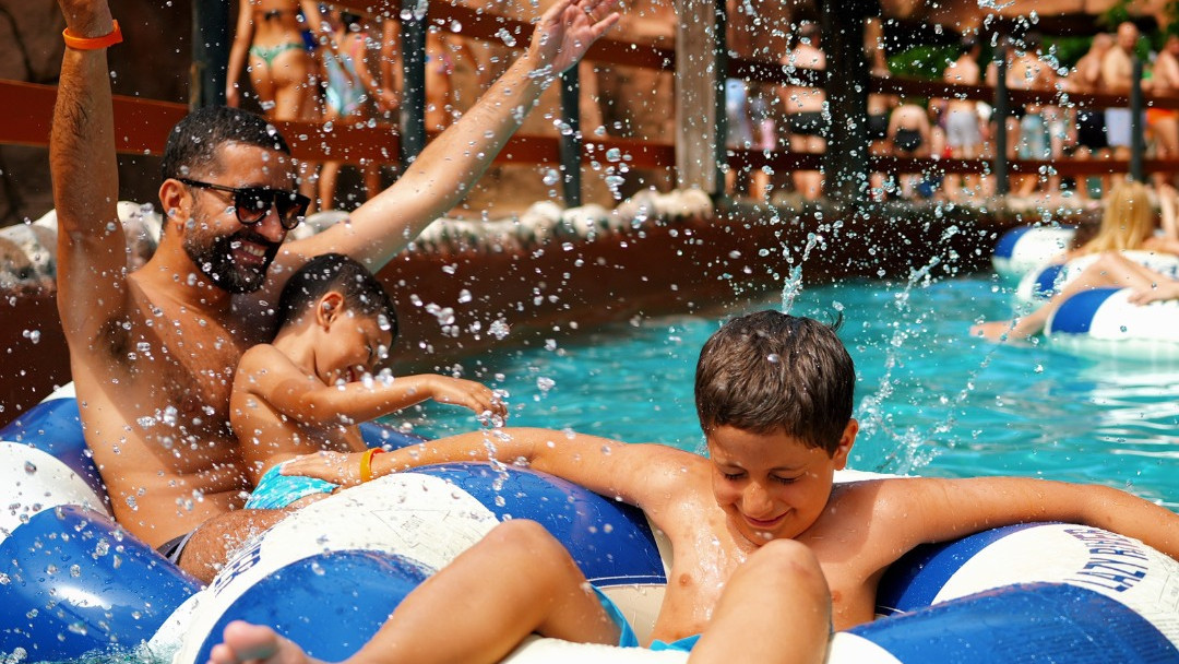 A man and his two young sons enjoying the Lazy River at Caneva Acquapark on a sunny day, laughing and splashing water while floating in inner tubes.