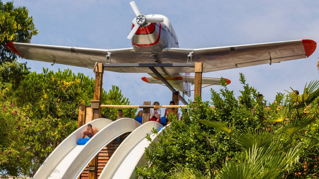 Guests racing down a white mat water slide at Caneva Acquapark, Italy, featuring a life-sized vintage propeller airplane as a themed entrance