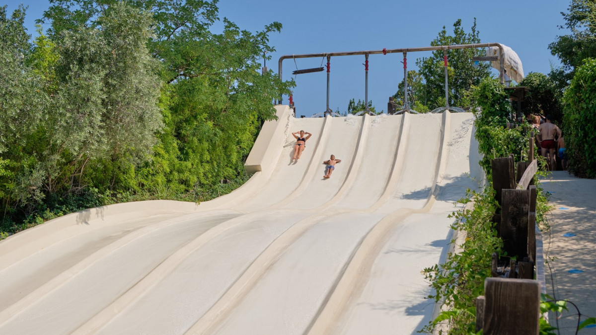 Two people racing down the multi-lane white water slide at Caneva Acquapark, surrounded by lush green trees on a clear summer day.