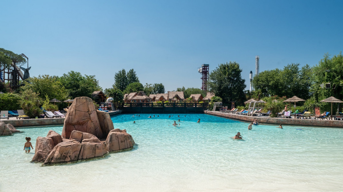 Wide view of the Shark Bay wave pool at Caneva Acquapark, featuring white sand entry, turquoise water with swimmers, and tropical palm trees under a clear blue sky