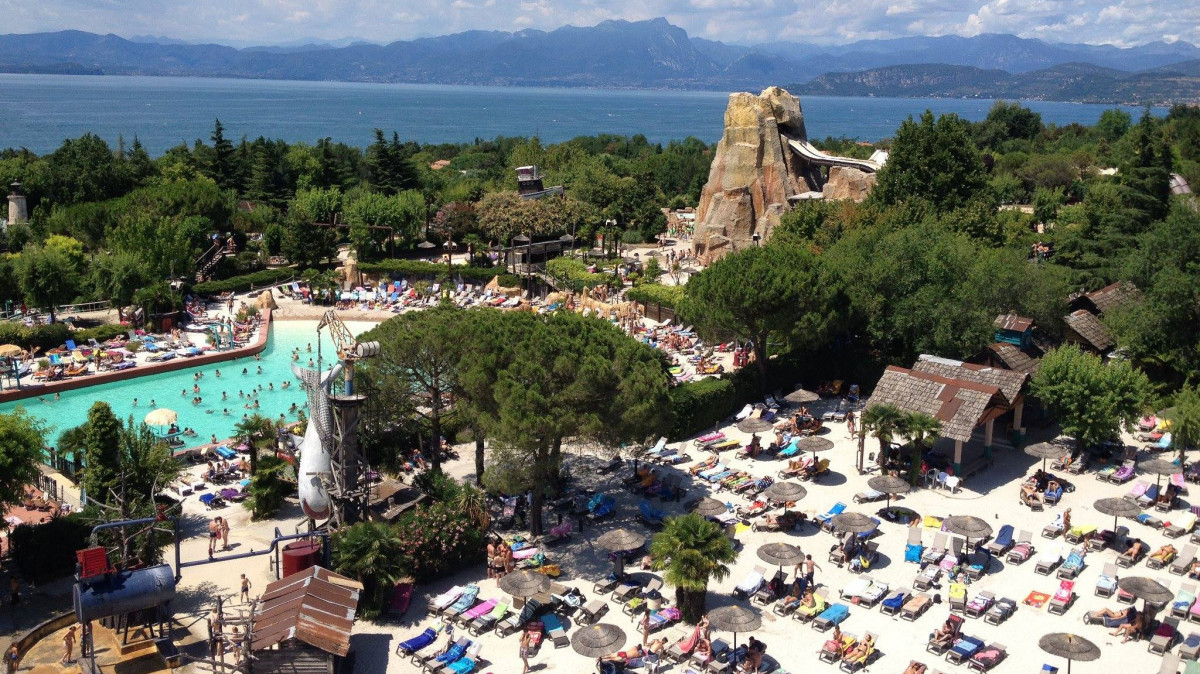 Breathtaking panoramic view of Caneva Acquapark in Italy, featuring tropical sandy beaches, swimming pools, and the iconic volcano water slide with Lake Garda and mountains in the background.
