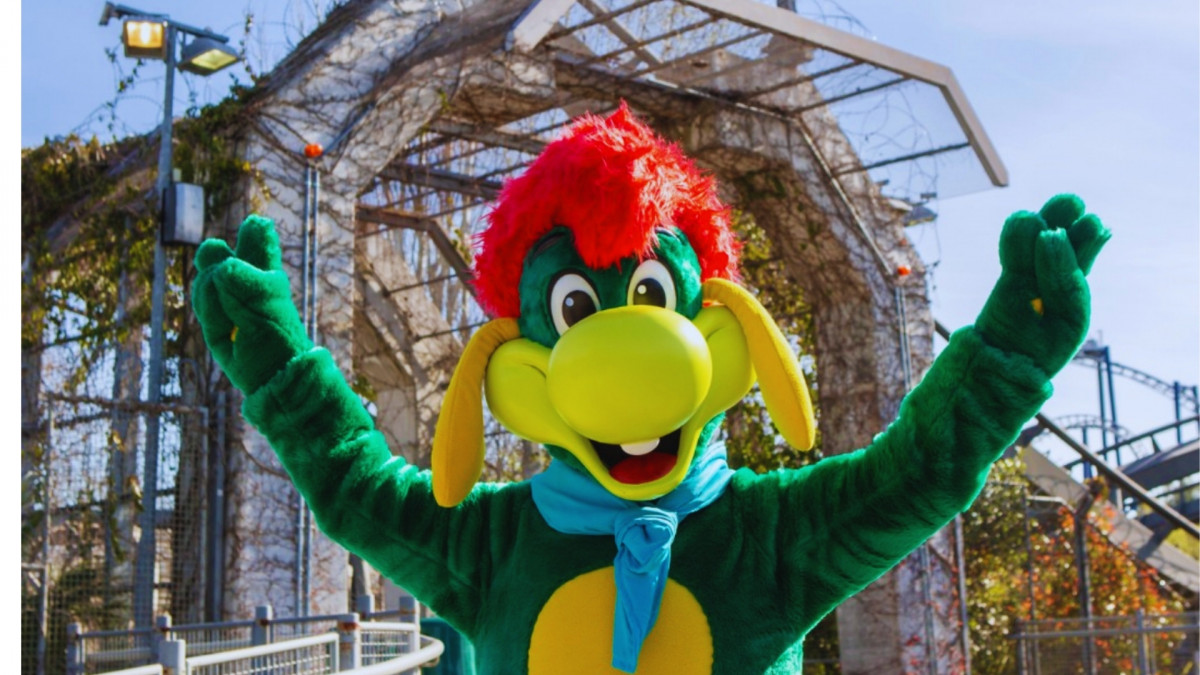 Prezzemolo mascot waving with Raptor roller coaster tracks in the background at Gardaland Park, Italy