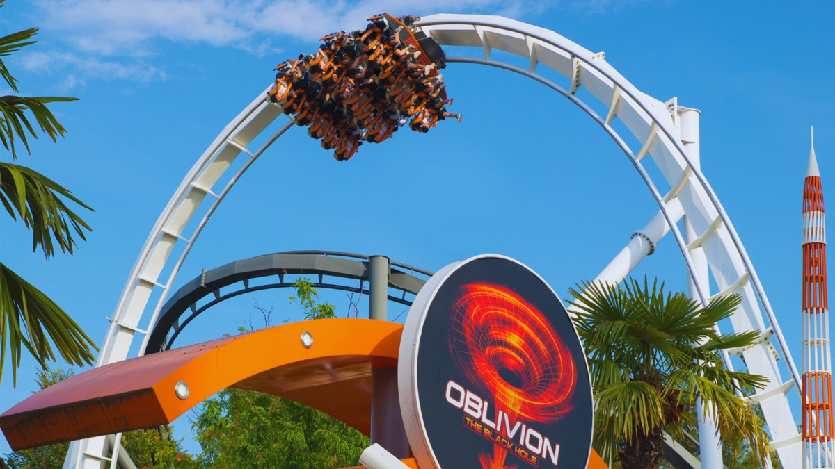The vertical drop of Oblivion dive coaster at Gardaland Park, Italy, with riders hanging over the edge against a blue sky.