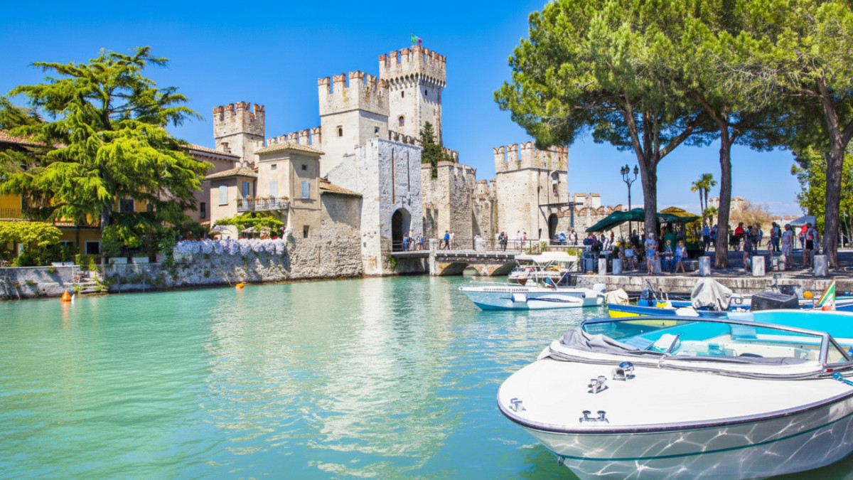 Scaligero Castle in Sirmione on the shores of Lake Garda, Italy, featuring a historic medieval fortress, a stone bridge, and boats moored in the turquoise water.