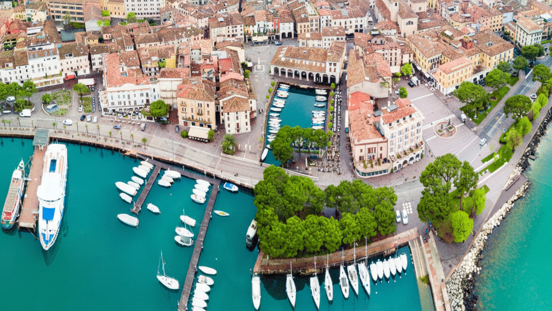 Aerial drone view of the historic town of Desenzano del Garda, Italy, featuring its scenic marina, docked boats, and traditional buildings on the shores of Lake Garda.