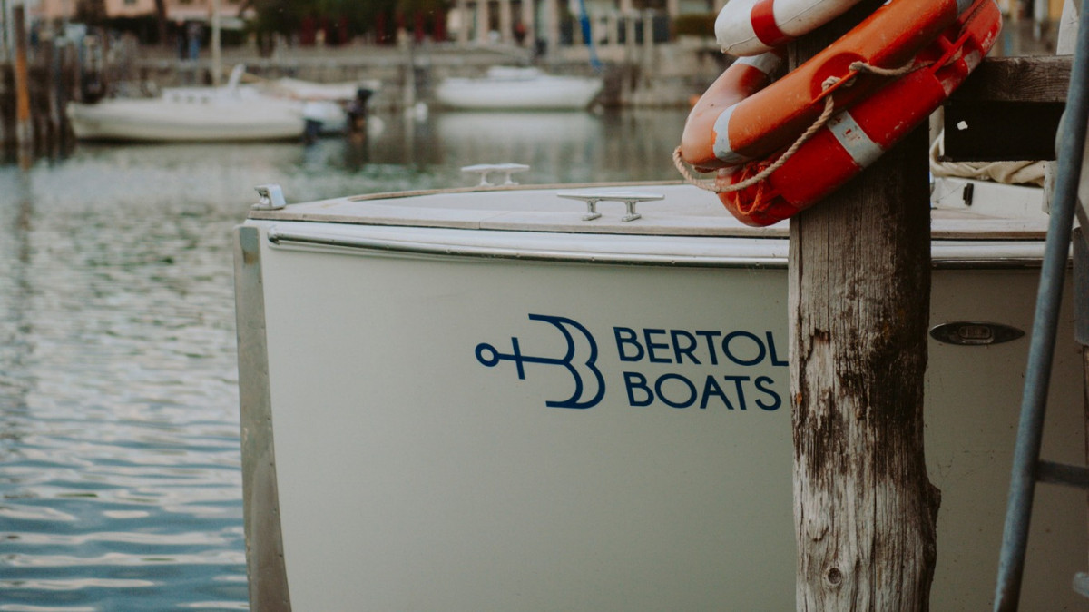 Side view of a Bertoldi Boats vessel docked at a pier, featuring the company logo on the hull and a lifebuoy on a wooden post.