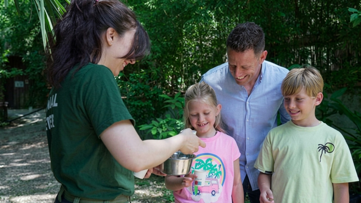 A smiling animal keeper is pouring a liquid from a stainless steel cup into a paper cup for a family during an outdoor educational activity