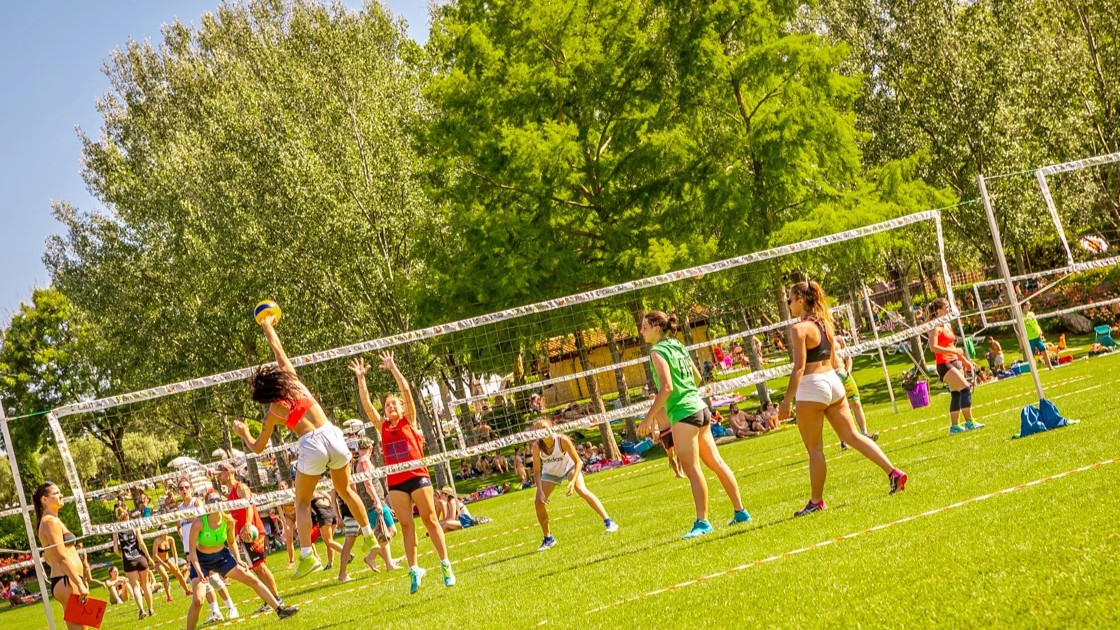 "A group of people playing beach volleyball on a green lawn at Cavour Waterpark, surrounded by lush trees under a sunny sky.