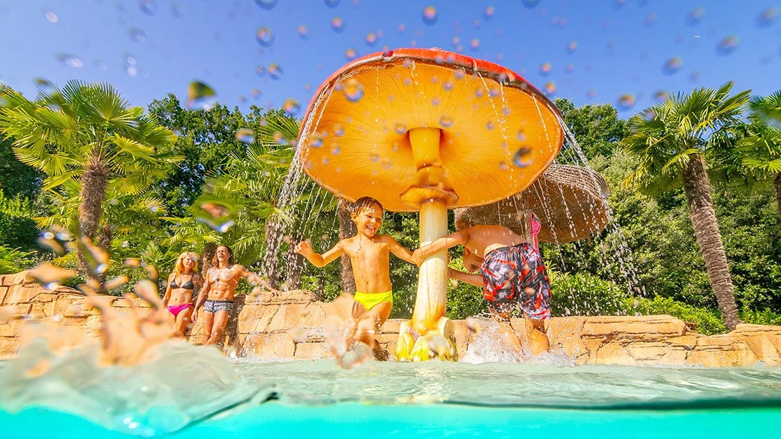 Children playing under a giant mushroom water fountain in a tropical-themed pool at Cavour Waterpark