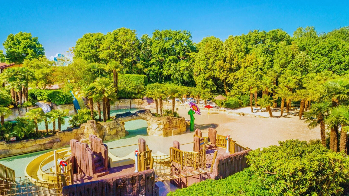 Tropical-themed water park area at Cavour Waterpark with artificial rock structures, palm trees, a shallow pool, and a white sand beach section under a clear blue sky