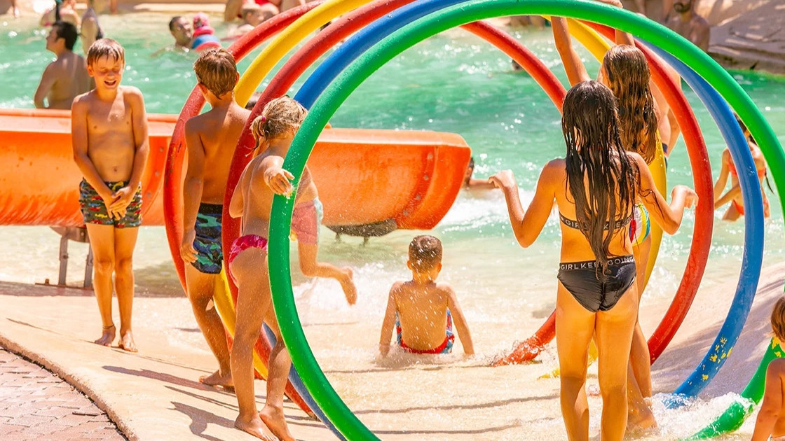 Children playing with colorful hoop water sprays and slides in a sunny outdoor pool area at Cavour Waterpark