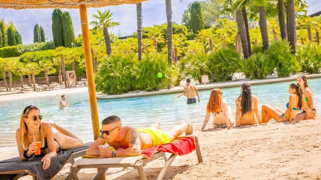 People relaxing on sunbeds and a sandy beach by a tropical pool at Cavour Waterpark, surrounded by palm trees