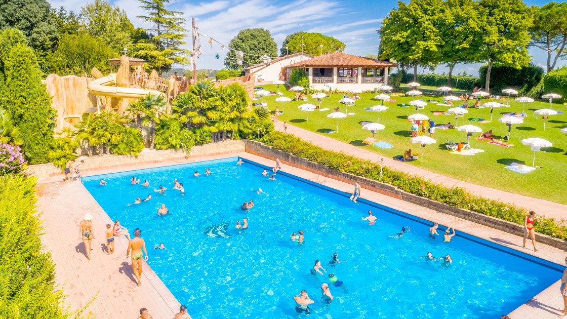 A large blue swimming pool with people, a yellow water slide, and a green lawn with white umbrellas at Cavour Waterpark