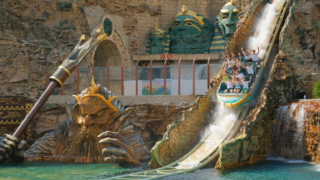 A thrilling splashdown of the Fuga da Atlantide water ride at Gardaland theme park in Italy, featuring a large mythological-themed boat with cheering riders and massive ancient statues.