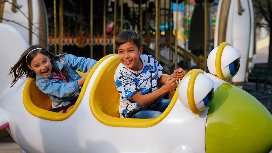 Two happy kids enjoying a kids' airplane ride at Gardaland theme park in Italy, with a carousel in the background.