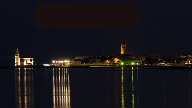Night skyline of Caorle, Italy, featuring the illuminated Sanctuary of Madonna dell'Angelo and bell tower reflecting on the sea.