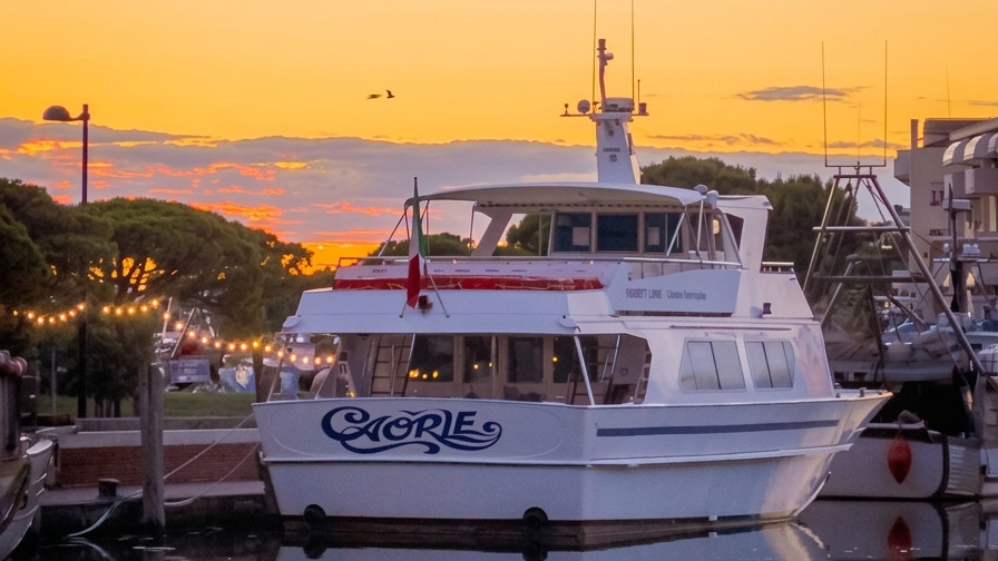 A close-up photograph of a white passenger motor yacht named "CAORLE" moored in a Italian marina at sunset, with string lights and trees in the background.