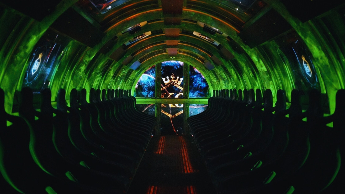 Interior of K6 The Royal Intelligence Agency interactive simulator ride at Movieland Park in Italy, featuring two rows of passenger seats within a futuristic circular pod under dynamic green lighting, with a screen showing the K6 agency logo.