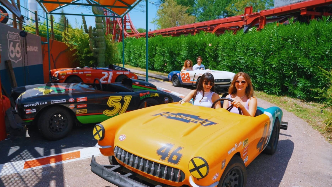Two young women smile while driving vintage-style go-karts  on the Route 66 attraction at Movieland Park in Italy