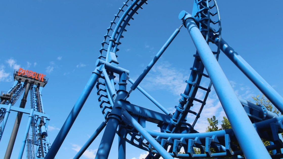 Low-angle photograph capturing the complex, vivid blue steel track and support structures of the Diabolik Invertigo rollercoaster at Movieland Park in Italy. On the far left, the red 'DIABOLIK' logo is visible on a tower, with a rollercoaster train ascend