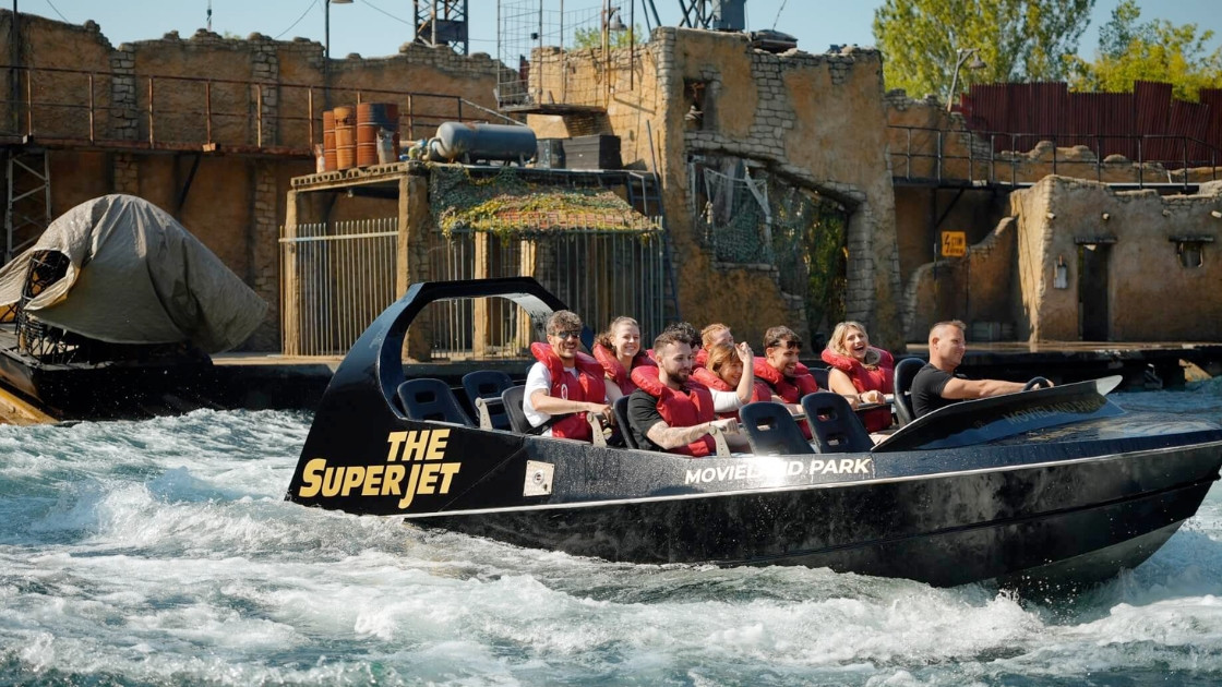 A group of people wearing red life jackets on The Superjet boat ride at Movieland Park, speeding through water with a themed stunt-set background.