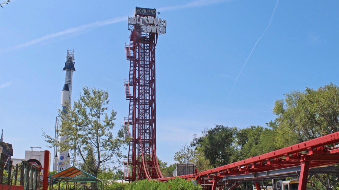 A tall, red Hollywood Action Tower drop ride at Movieland Park, Italy, with a large 'HOLLYWOOD TOWER' sign and a space launch rocket structure on the left against a blue sky with faint jet contrails.